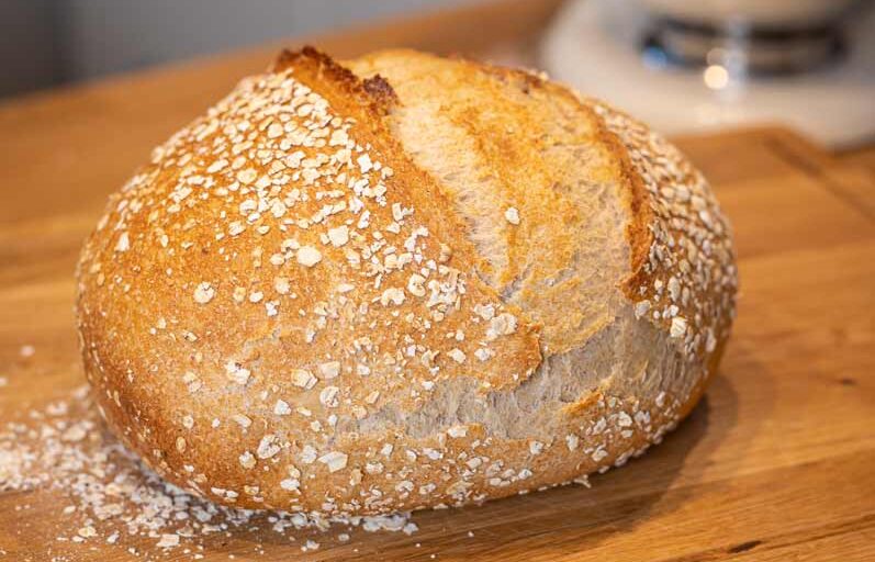 Laib Brot mit Haferflockenkruste auf einem Holztisch, im Hintergrund eine silberne Küchenmaschine.