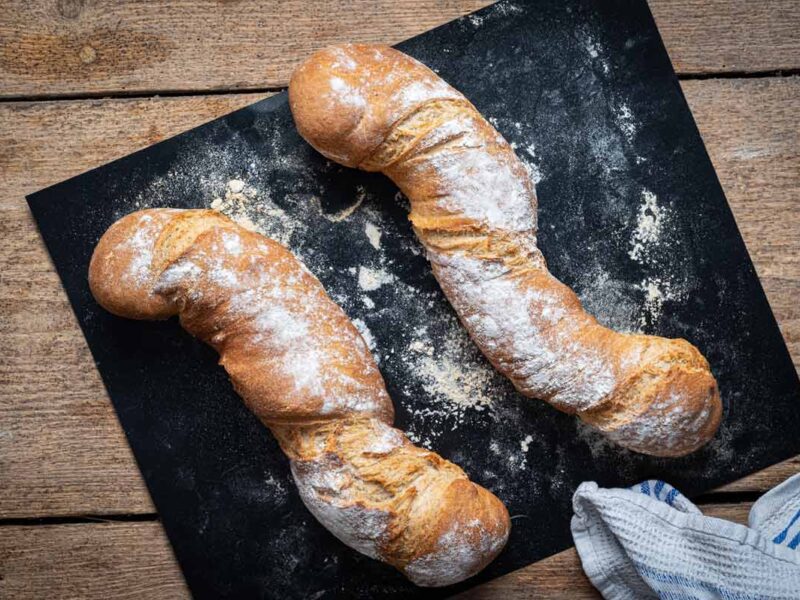 Zwei rustikale, längliche Brote auf schwarzem Backblech, bestäubt mit Mehl, auf Holztisch liegend.