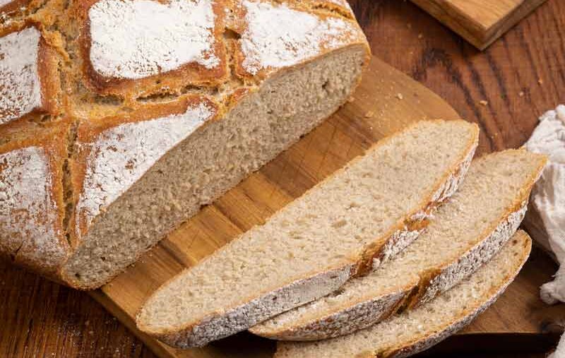 Frisch gebackenes, geschnittenes Brot auf einem Holzbrett, mit Brotmesser und Mehl auf einem Holztisch.