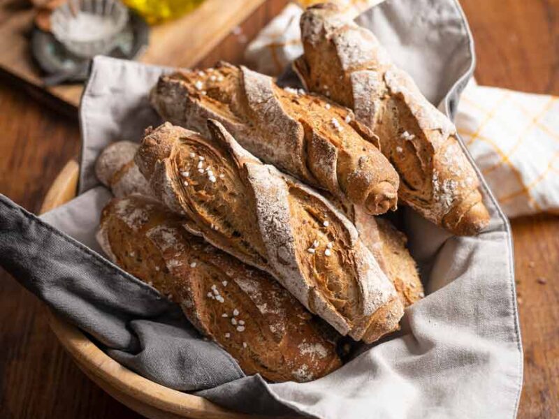 Frische Baguettes in einer Stoffserviette in einem Brotkorb, auf einem hölzernen Tisch, mit Ölflasche und Gewürzen im Hintergrund.
