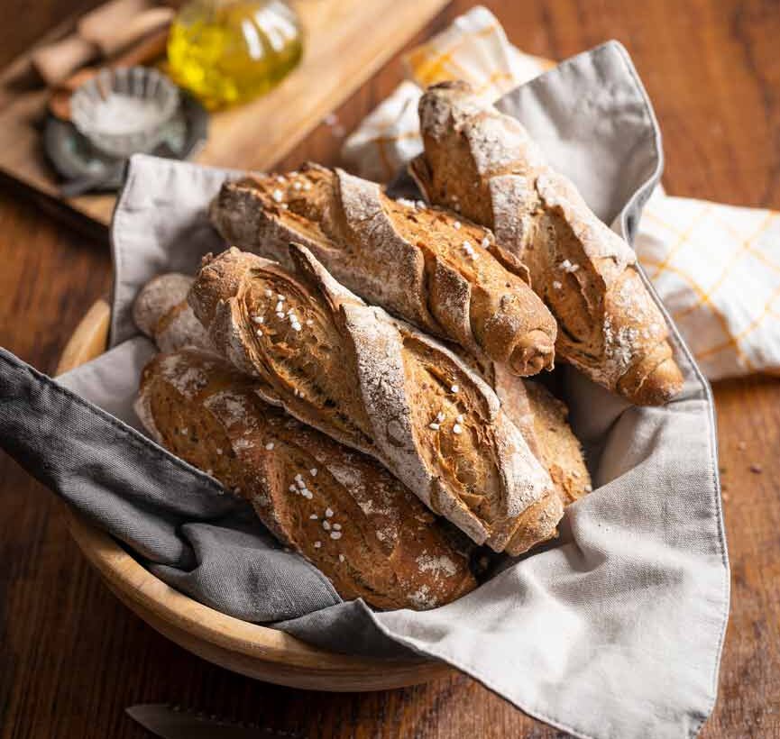 Eine Schale mit frischen, goldbraunen Baguettes auf einem Leinentuch, im Hintergrund ein Holzbrett mit Olivenöl.