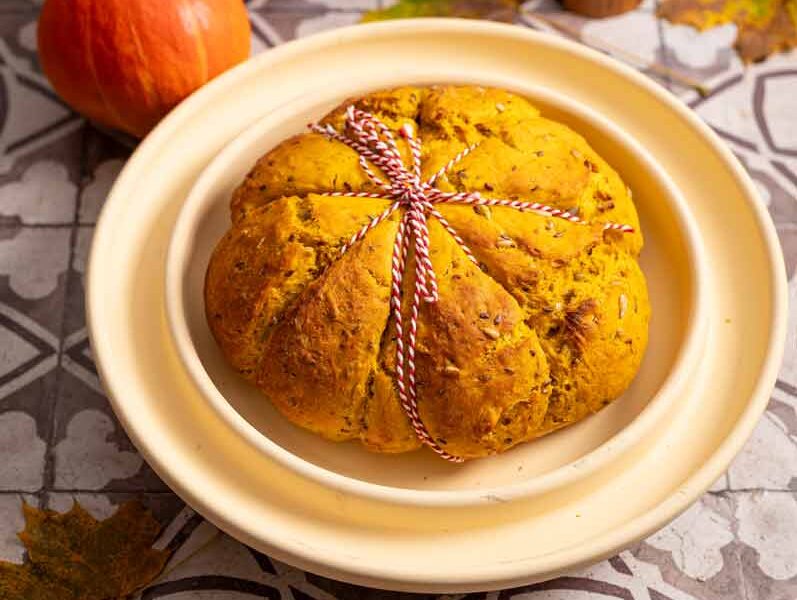 Kürbisförmiges Brot mit Schnur auf Teller, neben Kürbissen und Herbstblättern auf gemustertem Untergrund.