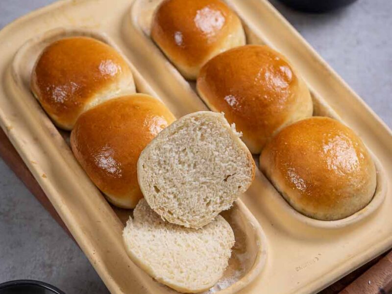 Vier goldbraune Brötchen in einer Form, eines ist aufgeschnitten. Im Hintergrund Mehl- und Milchschale.