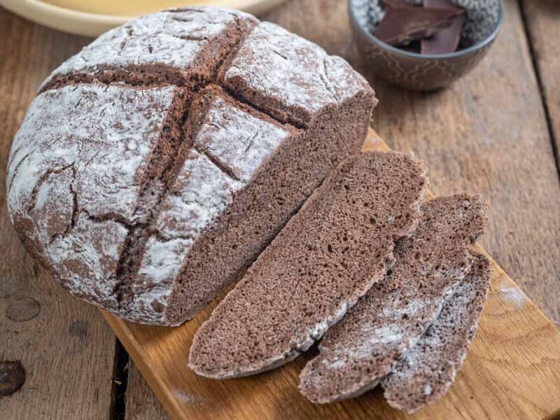 Leckeres Schokoladenbrot auf Holzbrett, teilweise geschnitten, mit Schokoladenstücken und Backutensilien im Hintergrund.