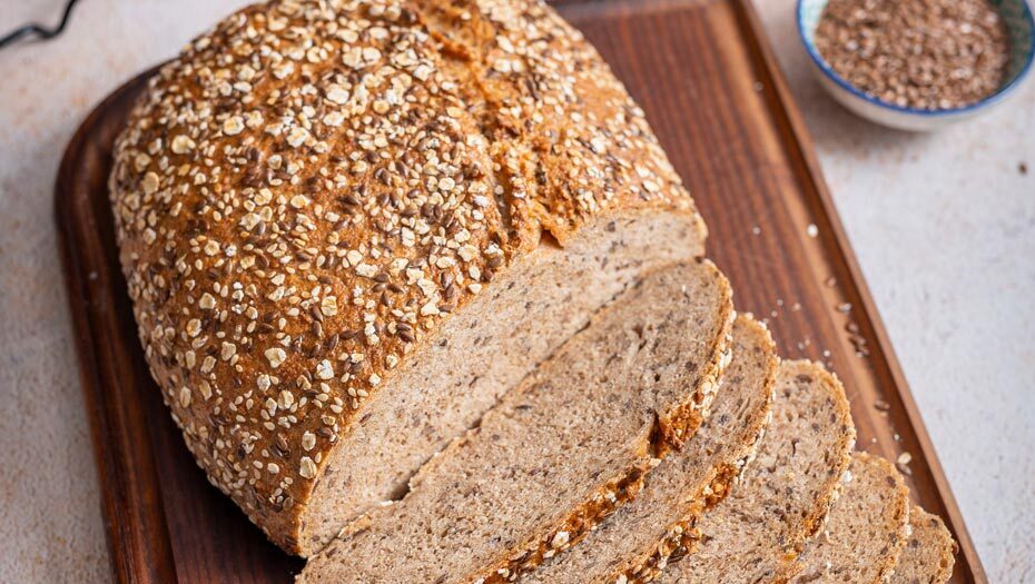 Ein geschnittenes Vollkornbrot mit Haferflocken auf einem hölzernen Schneidebrett, daneben ein Schälchen mit Saaten.