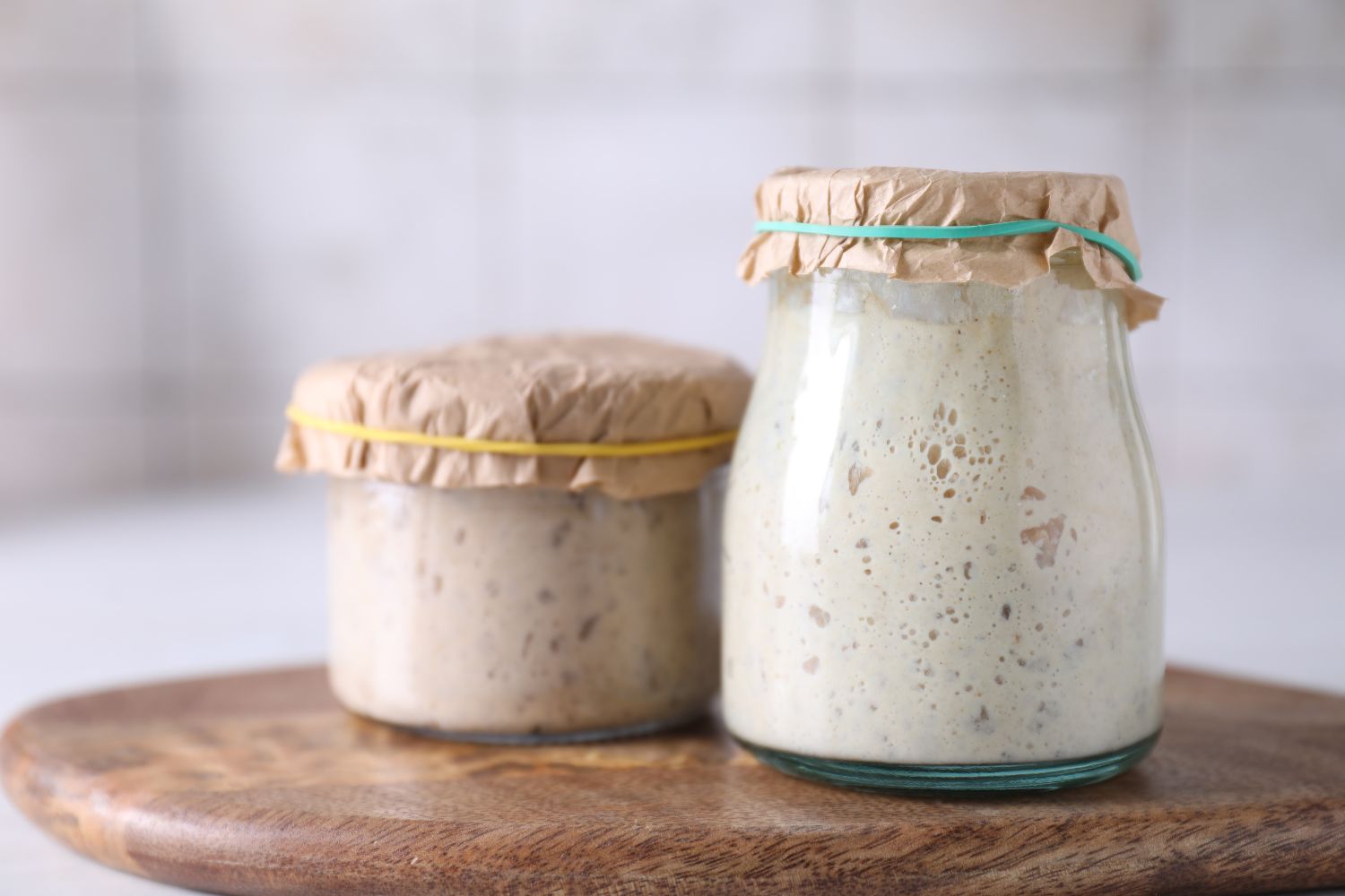 Zwei Gläser mit Sauerteig, abgedeckt mit Papier und Gummibändern, stehen auf einem Holzbrett vor Kachelhintergrund.