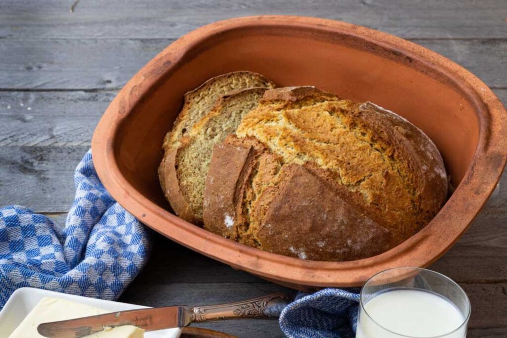 Frisch gebackenes Brot in einem Tontopf, daneben ein Glas Milch, ein Buttermesser und ein blau-weiß kariertes Tuch.