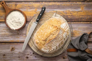 Laib Brot auf einem Gitter mit Messer, Holzschale mit Mehl und grauem Tuch auf rustikalem Holztisch.