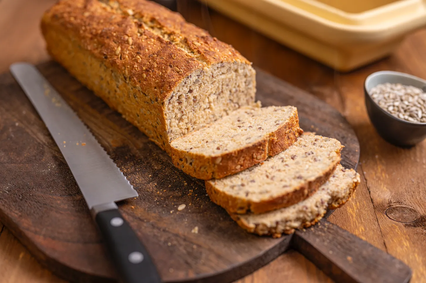 Mehrkornbrot auf Holzbrett, teilweise geschnitten, mit Brotmesser und Schälchen mit Sonnenblumenkernen im Hintergrund.