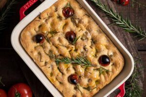 Focaccia in rotem Backform, mit Rosmarin, Kirschtomaten und Olivenöl, auf Holztisch mit frischem Rosmarin.