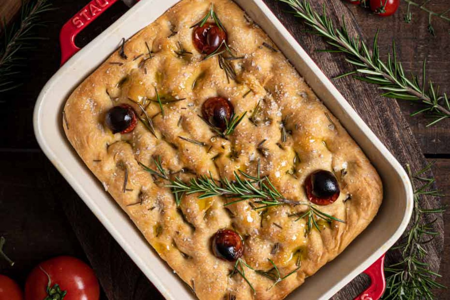Focaccia in rotem Backform, mit Rosmarin, Kirschtomaten und Olivenöl, auf Holztisch mit frischem Rosmarin.