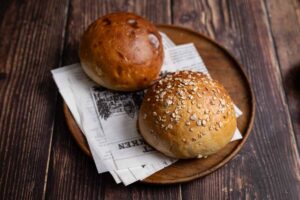 Zwei runde Brötchen, eins mit Haferflocken, auf Papierset, präsentiert auf einem Holzbrett mit dunklem Hintergrund.