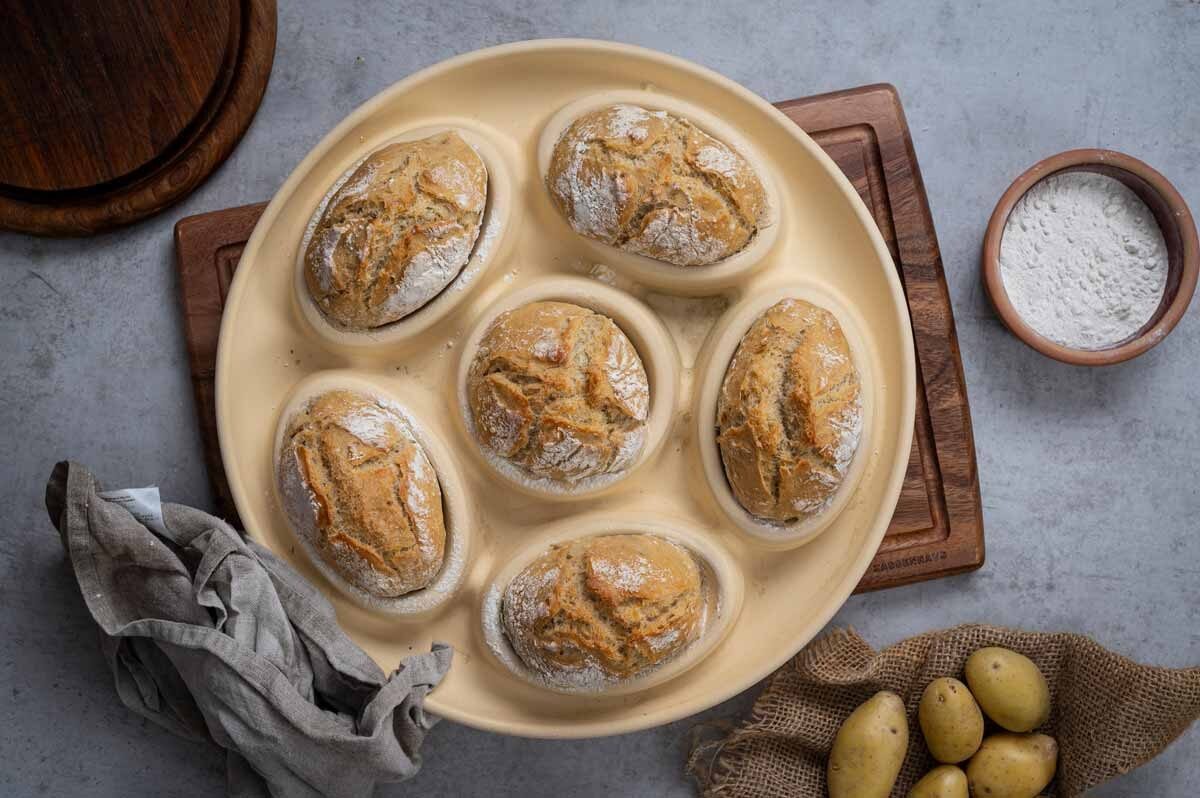 Sechs runde Brötchen in einer Backform, umgeben von einem Tuch, Kartoffeln und einer Schale mit Mehl.
