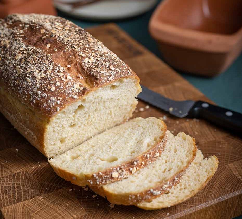 Ein frisch gebackenes Laib Brot mit Haferflocken, teilweise geschnitten, liegt auf einem Holzbrett neben einem Brotmesser.
