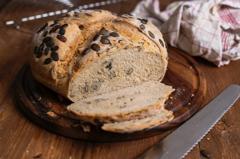 Laib Kürbiskernbrot auf Holzbrett, teilweise geschnitten, mit Brotmesser und Stofftuch im Hintergrund.