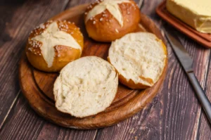 Drei Laugenbrötchen auf Holzbrett, eins aufgeschnitten. Messer und Butter im Hintergrund auf rustikalem Tisch.