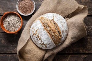 Rundes, rustikales Brot auf Sackleinen, dekoriert mit Mehlmotiv, daneben Schalen mit Leinsamen und Sonnenblumenkernen.