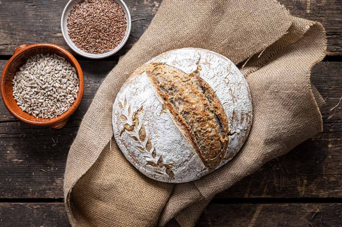 Rundes, rustikales Brot auf Sackleinen, dekoriert mit Mehlmotiv, daneben Schalen mit Leinsamen und Sonnenblumenkernen.