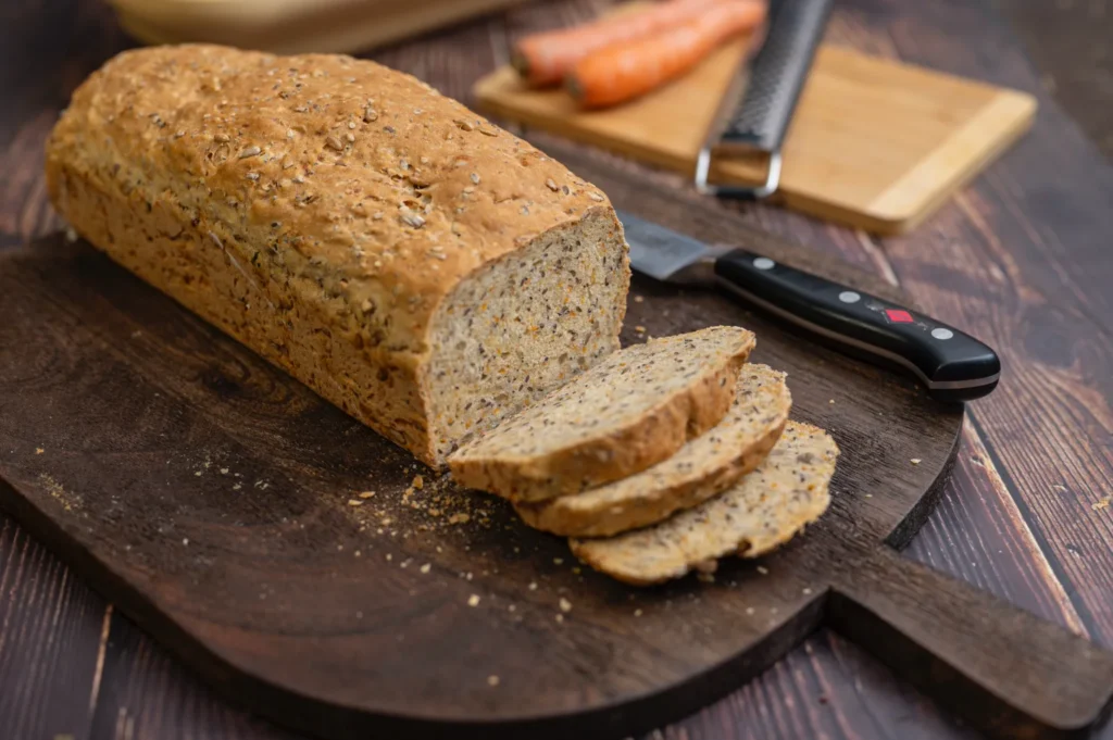 Ein Laib geschnittenes Vollkornbrot mit Körnern auf einem Holzbrett, daneben ein Messer und Karotten im Hintergrund.