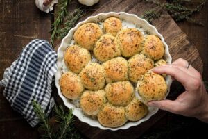 Hand nimmt ein mit Kräutern bestreutes, gebackenes Teigbrötchen aus einer runden, weißen Backform auf einem Holztisch.
