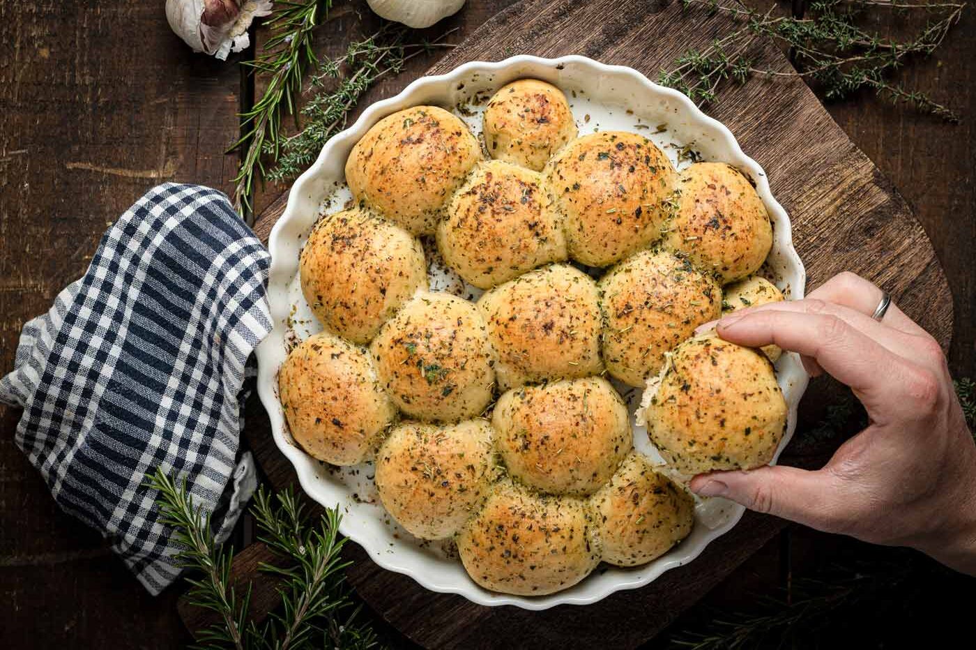 Hand nimmt ein mit Kräutern bestreutes, gebackenes Teigbrötchen aus einer runden, weißen Backform auf einem Holztisch.