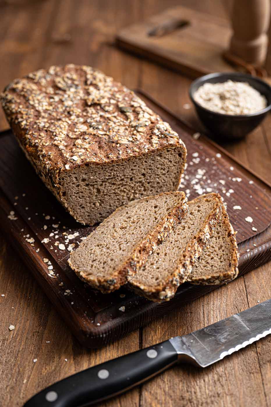 Vollkornbrot auf Holzbrett mit Haferflocken, zwei Scheiben abgeschnitten, daneben ein Messer und Schale mit Hafer.
