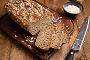 Ein geschnittenes Mehrkornbrot auf Holzbrett, umgeben von Haferflocken und Messer.