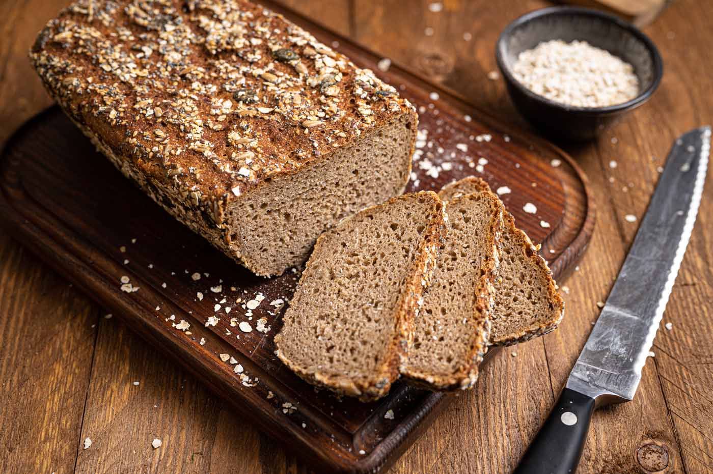 Ein geschnittenes Mehrkornbrot auf Holzbrett, umgeben von Haferflocken und Messer.