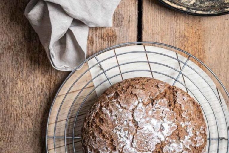 Rundes, frisch gebackenes Brot auf einem runden Kuchengitter auf Holztisch, daneben ein helles Tuch.