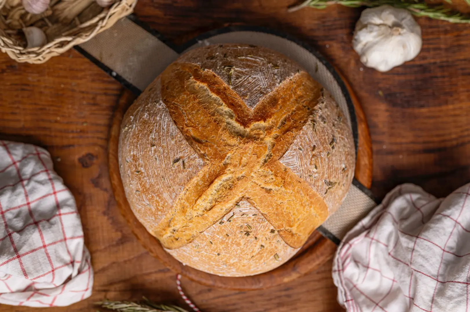 Rundes, goldenes Brot mit Kreuzmuster auf Holzbrett, umgeben von Knoblauchzehen, Tüchern und Rosmarinzweigen.