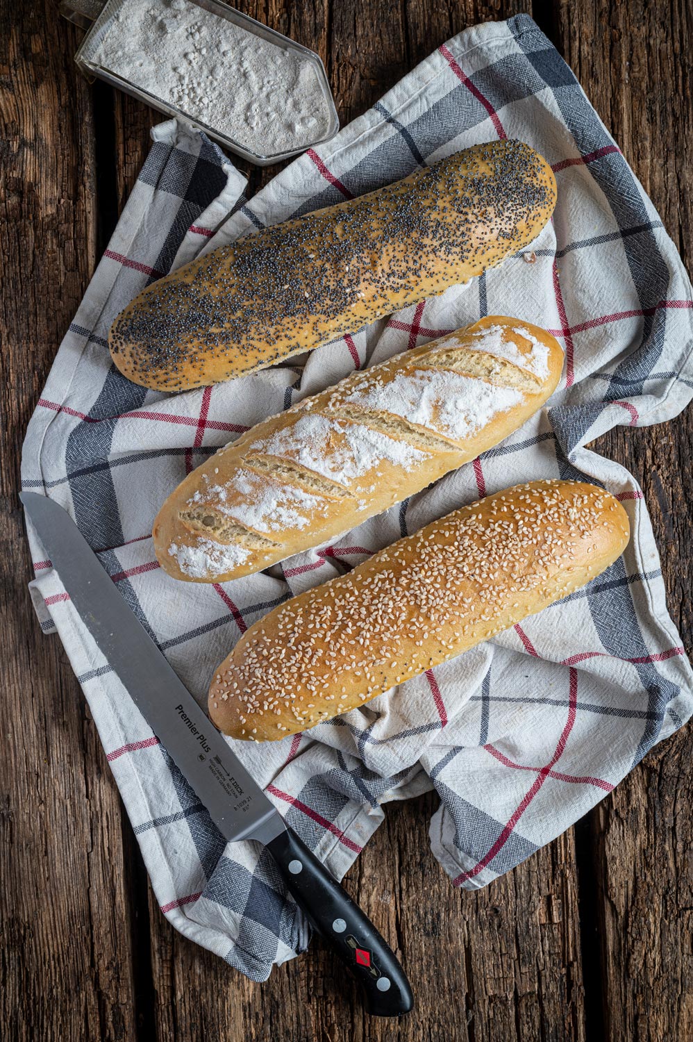 Drei Sandwich Baguettes mit Mohn, Mehl und Sesam auf einem karierten Küchentuch