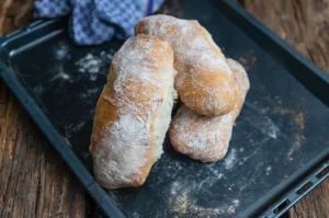 Drei frisch gebackene, bemehlte Brötchen auf einem schwarzen Backblech mit blauem Tuch im Hintergrund.