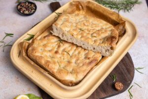 Frische Focaccia mit Rosmarin in gelber Backform, daneben Pfefferkörner und Zitrone auf Holzbrett.