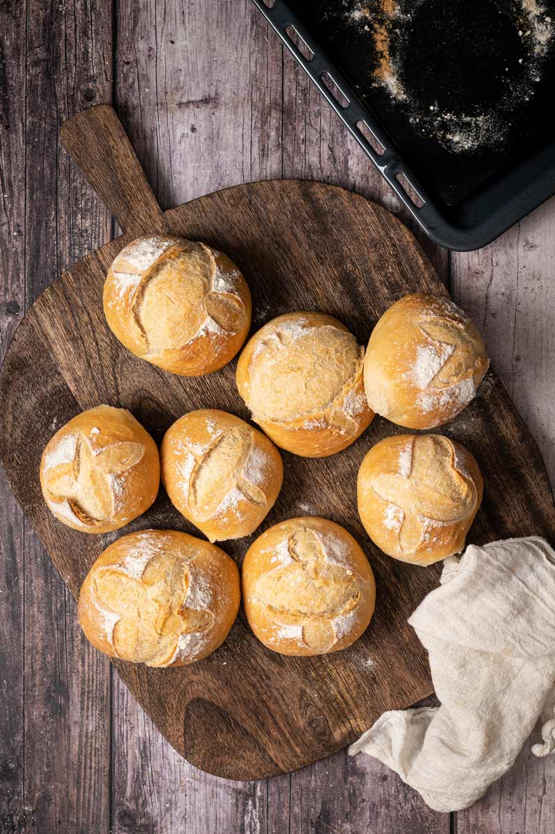 Acht Brötchen auf einem hölzernen Schneidebrett, daneben ein Geschirrtuch und ein Backblech im Hintergrund.