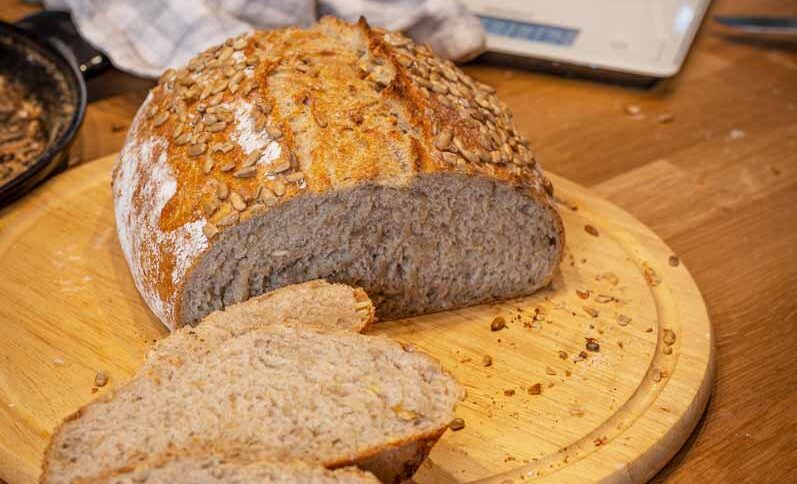 Brotlaib mit Körnerkruste, teilweise geschnitten, liegt auf einem runden Holzbrett auf einem Tisch.