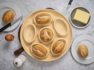 Sechs Brötchen in einer runden, hellen Backform, umgeben von Ei, Butter, Messer und Pfeffermühle auf Marmortisch.
