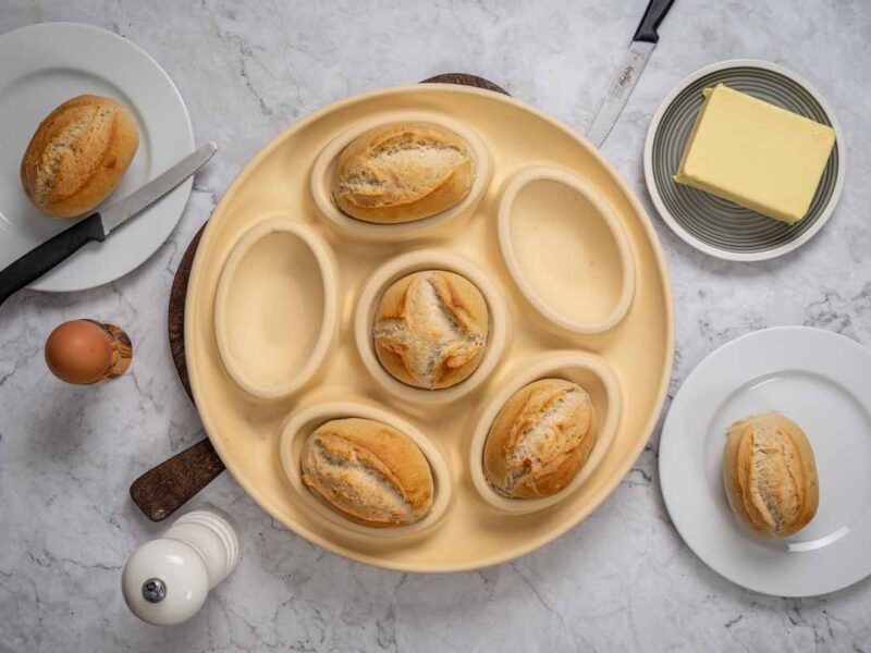 Sechs Brötchen in einer runden, hellen Backform, umgeben von Ei, Butter, Messer und Pfeffermühle auf Marmortisch.