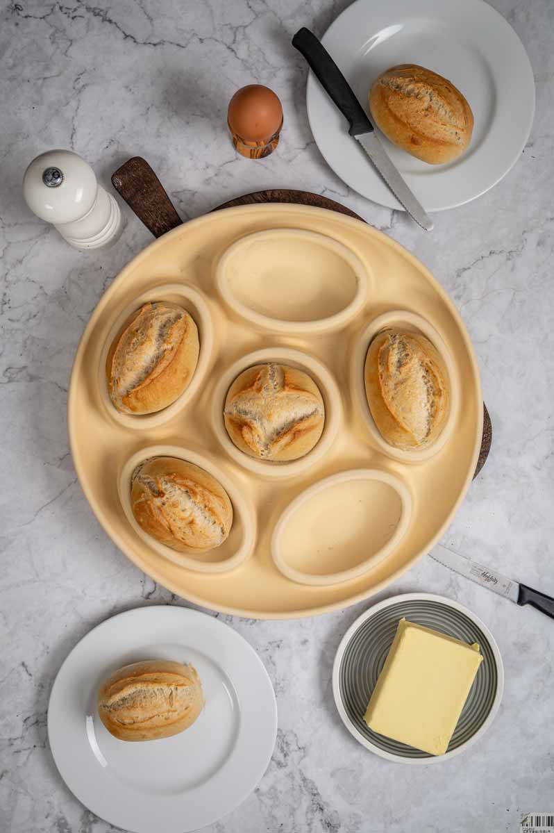Runde Backform mit frischen Brötchen auf Marmortisch. Ein Ei im Becher, ein Messer, Teller mit Brötchen, Butterschale dabei.