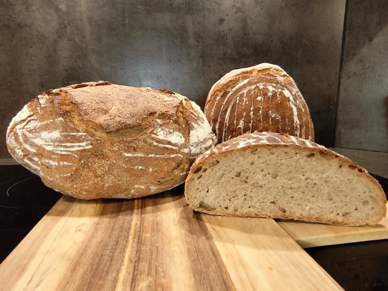 Drei Laibe frisches Brot, eins davon aufgeschnitten, liegen auf einem hölzernen Schneidebrett in einer Küche.