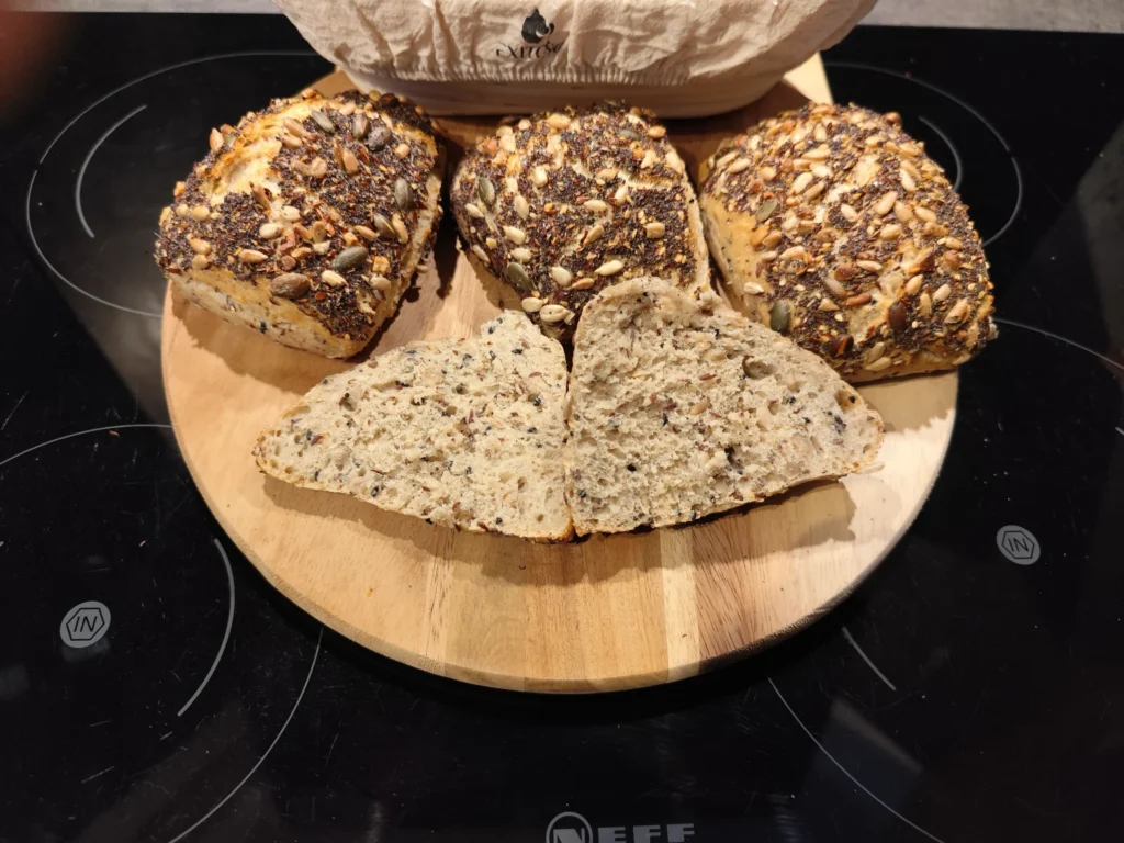 Fünf Mehrkornbrötchen mit Samen auf einem runden Holzbrett, eins aufgeschnitten, zeigen Körner im Inneren.