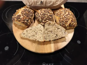 Fünf Mehrkornbrötchen mit Samen auf einem runden Holzbrett, eins aufgeschnitten, zeigen Körner im Inneren.
