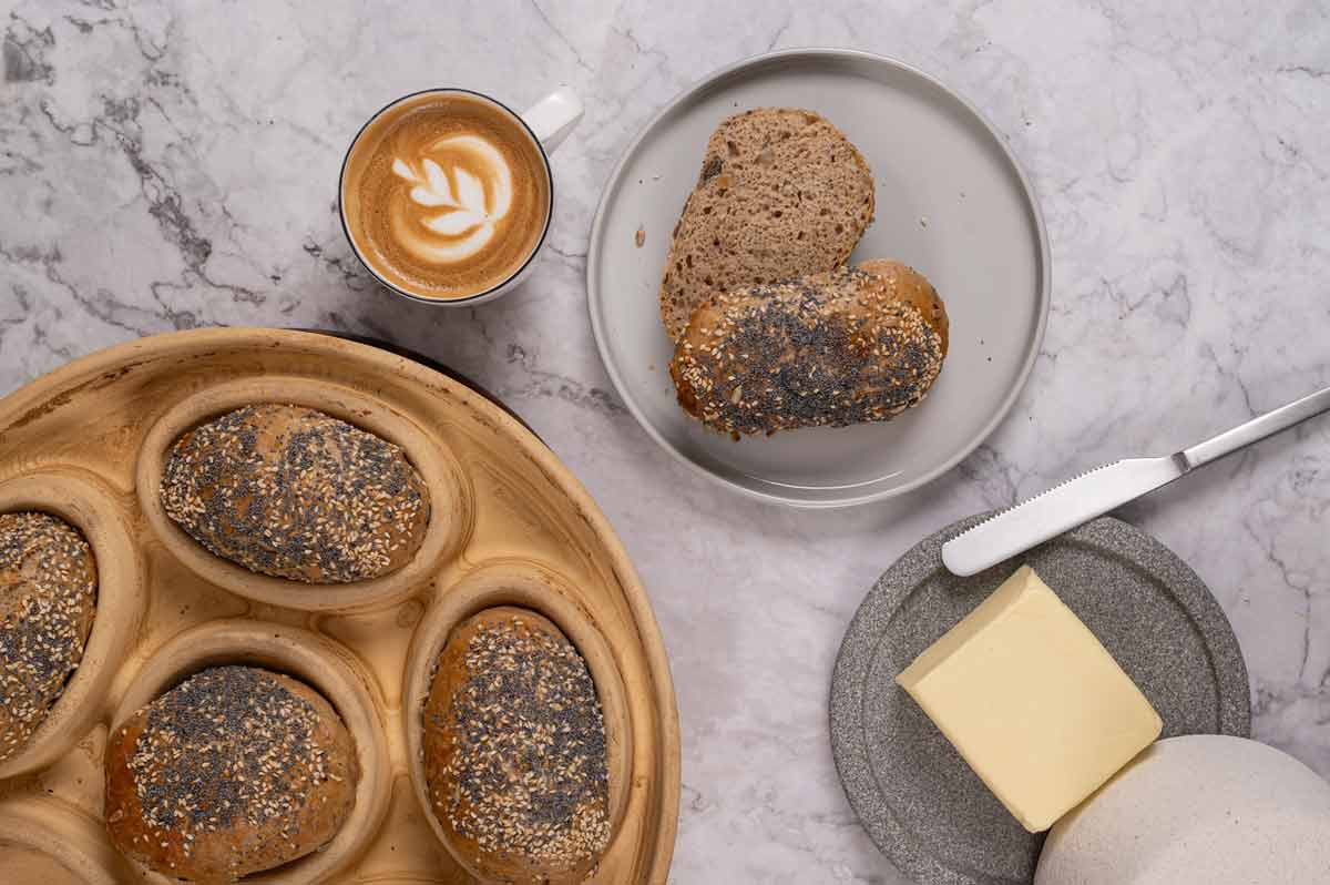 Teller mit Mohnbrötchen, daneben ein Cappuccino, ein Messer und ein Butterstück auf einem grauen Tisch.