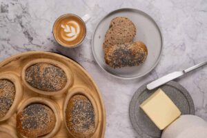 Frühstücksszene mit Mohn- und Sesambrötchen, geschnittenem Brötchen auf Teller, Kaffee mit Latte Art und Butterstück auf Marmortisch.