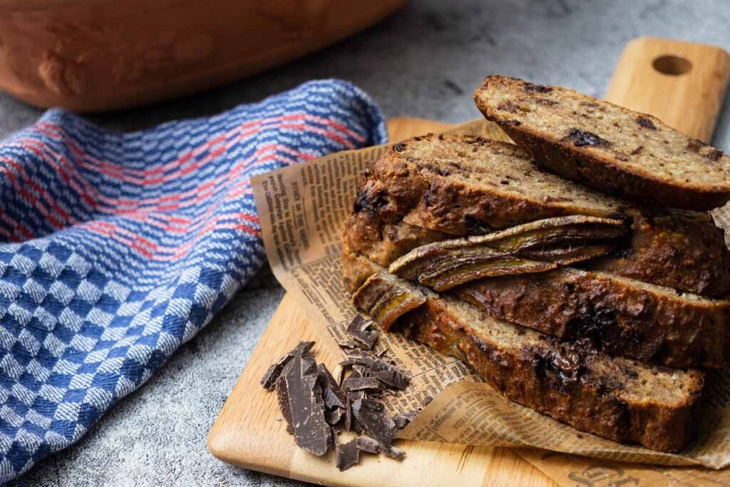 Bananenbrot-Scheiben auf Holzbrett, daneben Zartbitterschokoladenstücke und blau kariertes Tuch.