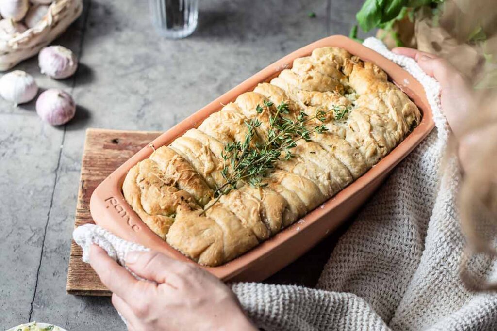 Hände halten eine Terrakotta-Backform mit frischem Brot, garniert mit Thymianzweigen, auf einem Holzbrett.