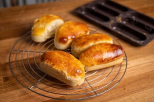 Fünf goldbraune, frisch gebackene Brötchen auf einem Gitterrost auf einem Holztisch. Im Hintergrund steht eine leere Backform.