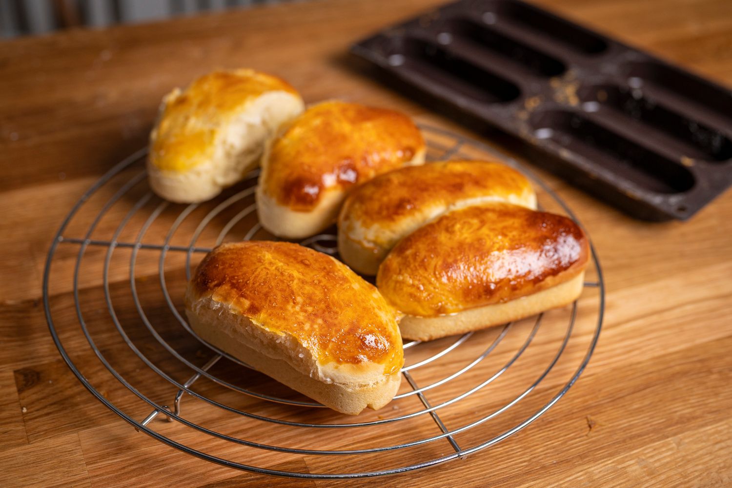 Fünf goldbraune, frisch gebackene Brötchen auf einem Gitterrost auf einem Holztisch. Im Hintergrund steht eine leere Backform.