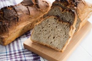 Ein Laib Brot und mehrere Brotscheiben auf einem Holzbrett, daneben ein kariertes Tuch.
