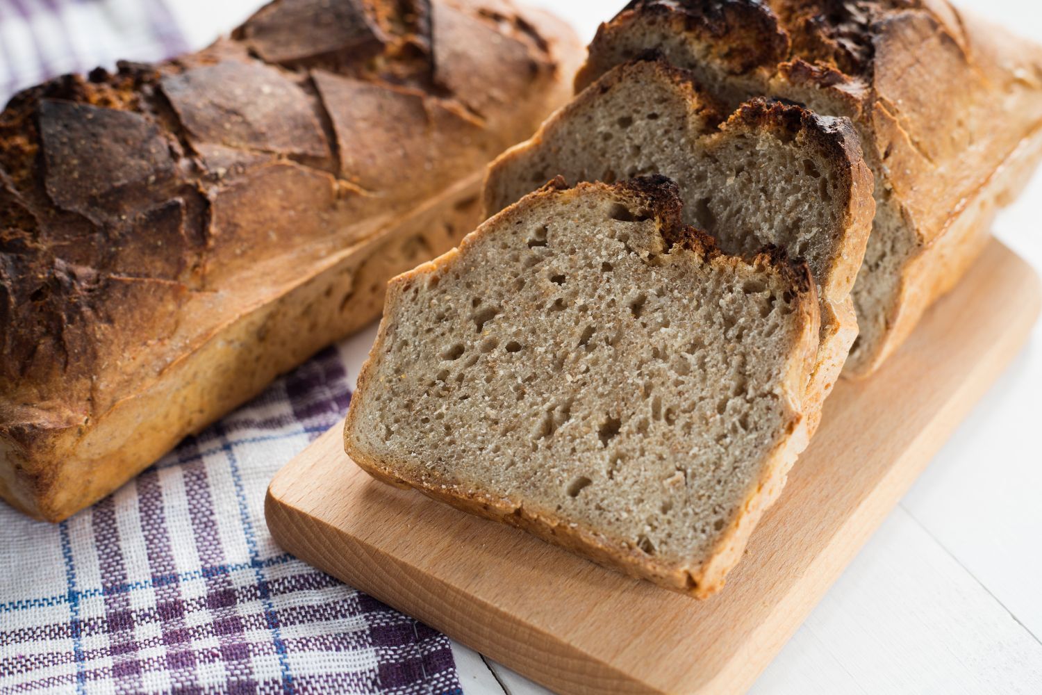 Ein Laib Brot und mehrere Brotscheiben auf einem Holzbrett, daneben ein kariertes Tuch.