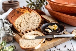 Ein angeschnittenes Brot auf einem Holzbrett, mit Buttermesser, Butter und Gewürzglas in der Nähe.